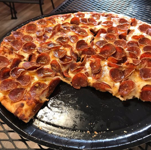 A large pepperoni pizza on a black tray, with a slice missing and toppings evenly distributed.