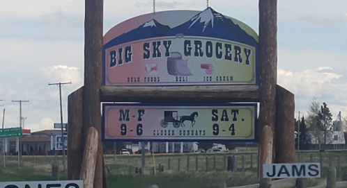 Sign for Big Sky Grocery featuring hours: M-F 9-6, Sat 9-4, closed Sundays, with mountains in the background.