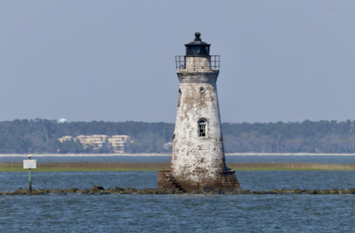 A weathered lighthouse stands on a rocky outcrop in calm waters, with trees and buildings in the background.