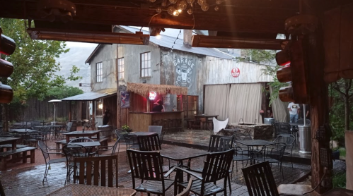 A rustic outdoor dining area with metal tables and chairs, surrounded by a wooden structure, under rainy weather.