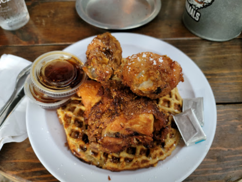 A plate featuring fried chicken on a waffle, drizzled with syrup, and accompanied by butter packets.