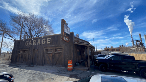 A rustic garage building with a sign, surrounded by industrial structures and a blue sky with clouds.