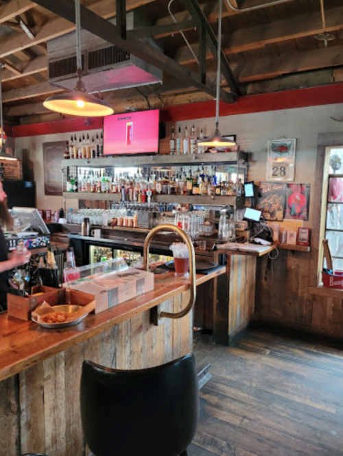 A cozy bar interior with a wooden counter, shelves of liquor, and a TV screen, featuring a rustic atmosphere.