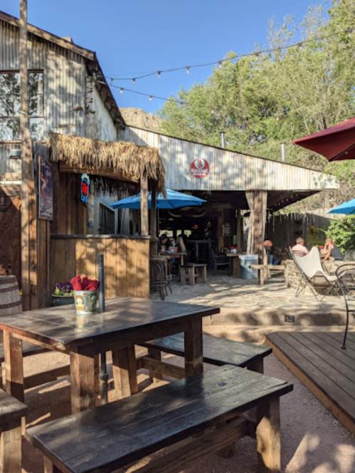 Outdoor seating area of a rustic bar with wooden tables, umbrellas, and a relaxed atmosphere surrounded by greenery.