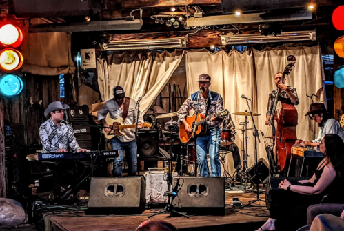 A live band performs on stage with instruments, including guitar and keyboard, in a rustic venue. Audience members watch.