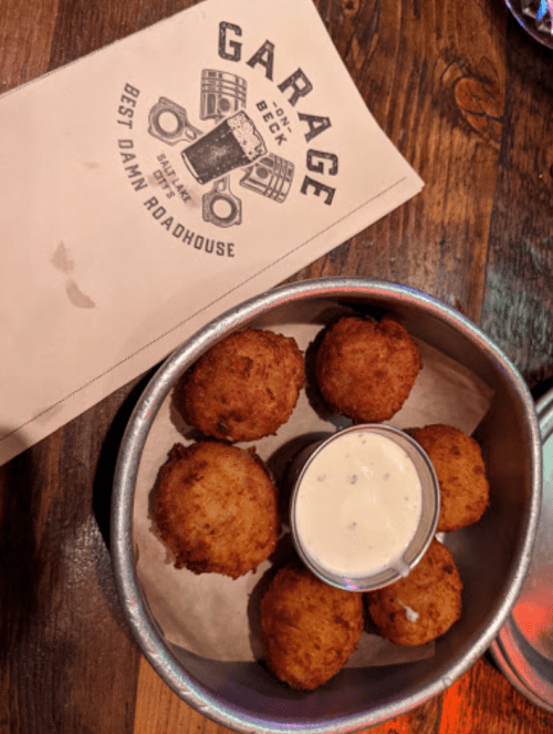 A metal bowl with six golden-brown fritters and a small cup of dipping sauce, placed on a wooden table with a menu.