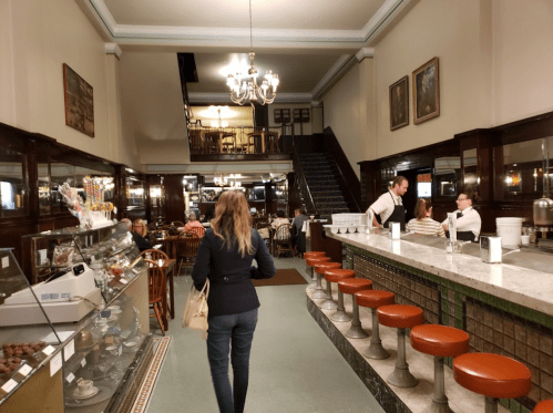 A woman walks through a vintage café with wooden decor, a counter, and patrons seated at tables.