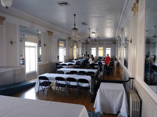 A spacious room with tables and chairs set up for an event, featuring large windows and elegant decor.