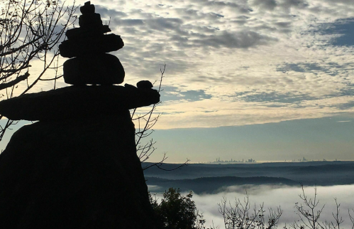 A silhouette of stacked stones against a cloudy sky, overlooking misty hills and a distant city skyline.