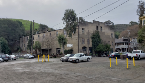 A gravel parking lot with several cars in front of a large, weathered building surrounded by greenery and hills.