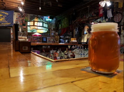 A jar of beer sits on a wooden bar, with a lively pub atmosphere and various decorations in the background.
