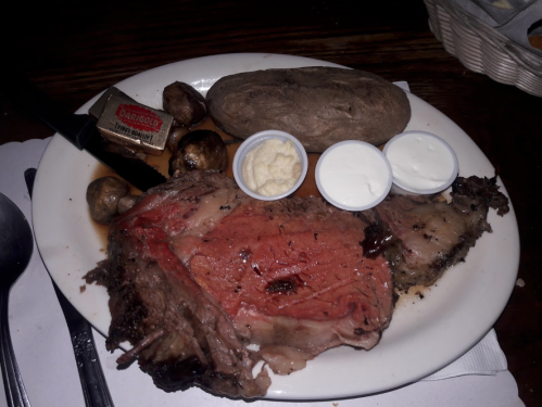 A plate with a slice of prime rib, baked potato, mushrooms, and two dipping sauces.