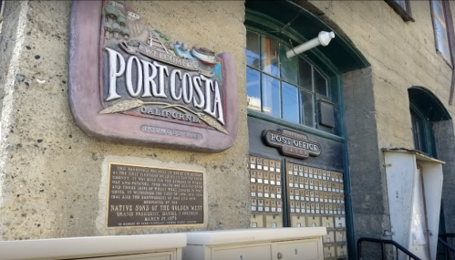 Sign for Port Costa, California, with a historic plaque and a post office entrance visible on a stone wall.