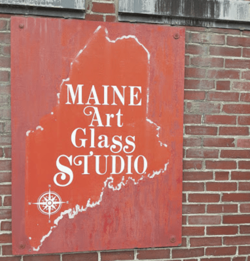 Sign for "Maine Art Glass Studio" featuring a map outline of Maine on a red background against a brick wall.