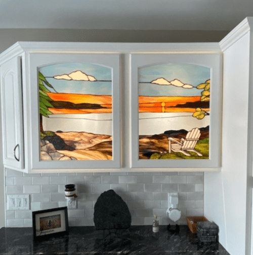 Stained glass windows depicting a sunset over water, with a tree and an Adirondack chair, framed in white cabinetry.