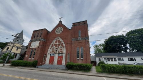 A red brick church with a cross on top, featuring large arched windows and a staircase, set against a cloudy sky.