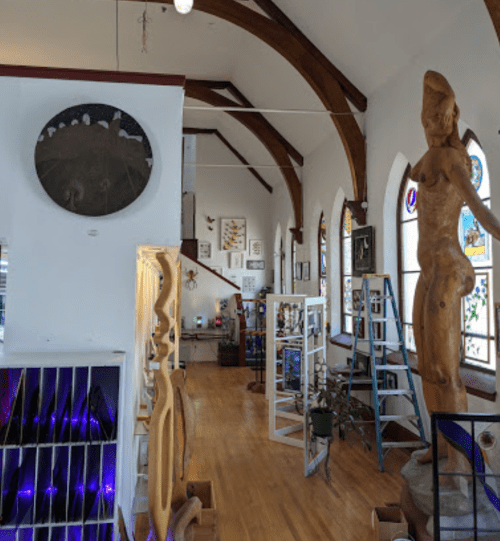 Interior of an art gallery featuring wooden sculptures, stained glass windows, and a high ceiling with wooden beams.