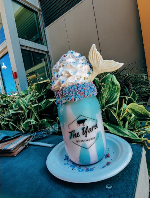 A colorful milkshake topped with whipped cream, sprinkles, and a mermaid tail, served in a jar on a plate.