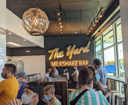 A busy milkshake bar interior with a neon sign reading "The Yard Milkshake Bar" and customers waiting in line.