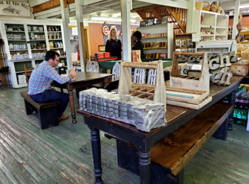 A man sits at a table in a market, surrounded by wooden displays and shelves filled with various products.