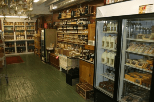 A cozy store interior filled with shelves of jars, bottles, and packaged goods, with a fridge displaying beverages.