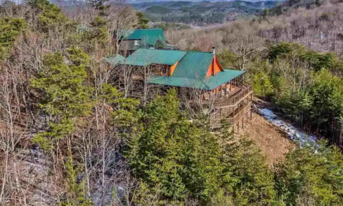 Aerial view of a cabin with green roofs nestled among trees on a hillside, surrounded by mountains.