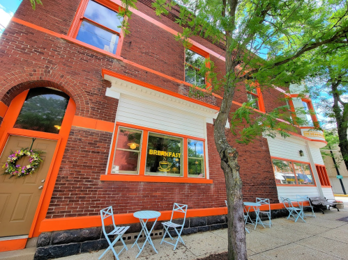 A brick building with orange accents, featuring a "Breakfast" sign and outdoor seating under a tree.