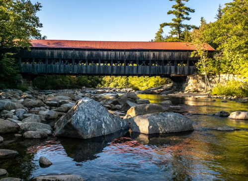 A covered bridge spans a river, surrounded by trees and large rocks, under a clear blue sky.