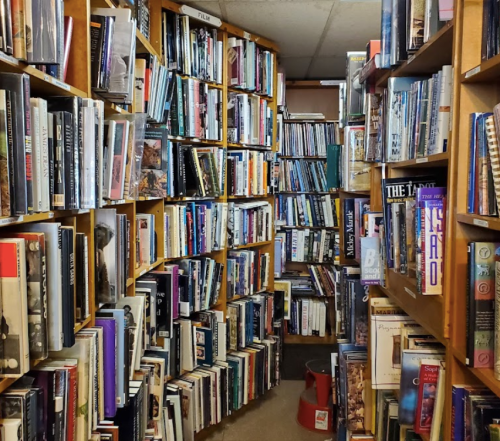 A narrow aisle between tall bookshelves filled with various books in a cozy, cluttered library.
