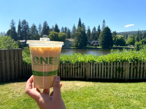 A hand holds a drink cup in front of a serene lake and lush greenery under a clear blue sky.