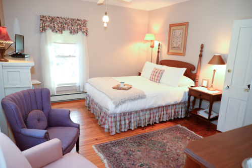 Cozy bedroom with a queen bed, armchair, wooden furniture, and warm lighting, featuring a rug and decorative curtains.
