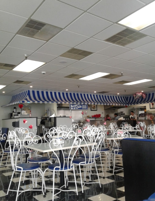 A bright, empty diner with blue and white striped awning, featuring white tables and chairs, and a checkered floor.