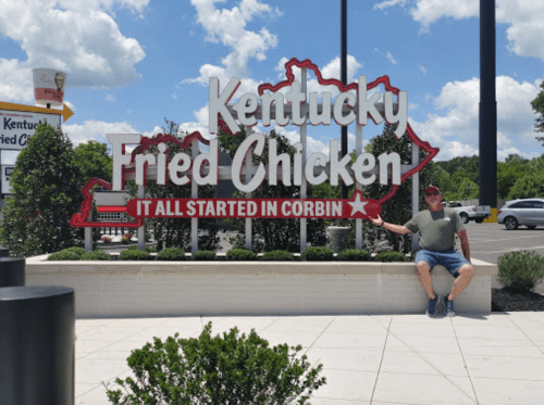 A person sits next to a large sign for Kentucky Fried Chicken, stating "It all started in Corbin." Bright, sunny day.