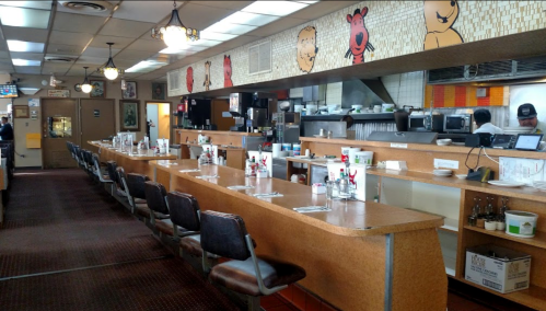 A diner interior featuring a long counter with stools, colorful wall art, and a kitchen area in the background.