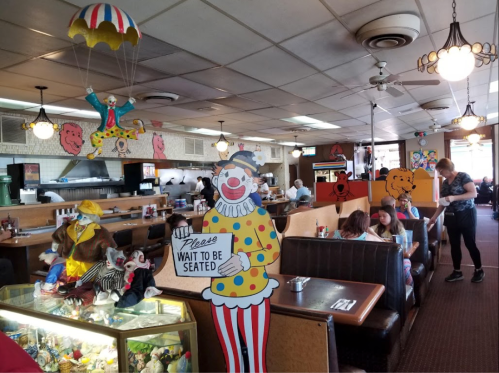 A colorful diner interior with clown decorations, families dining, and a sign reading "Please wait to be seated."