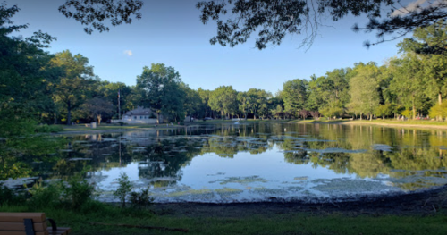 A serene lake surrounded by trees, reflecting the sky and greenery, with a small building in the background.