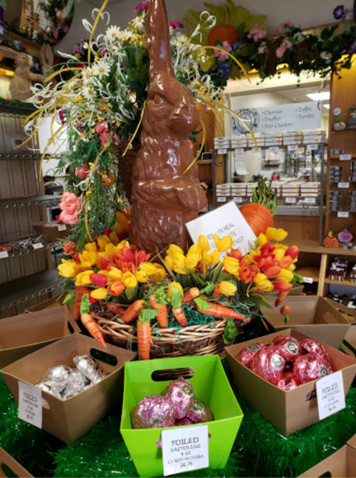 A large chocolate bunny surrounded by colorful flowers and Easter eggs in baskets on a festive display.