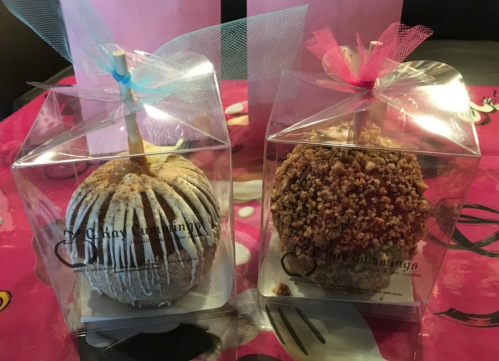 Two decorative caramel apples in clear boxes, one drizzled with white chocolate and the other coated in nuts.