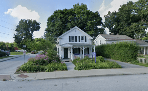 A charming white house with a porch, surrounded by greenery and flowers, on a quiet street.