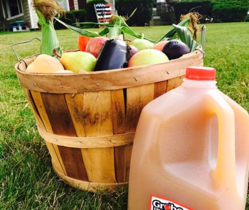 A wooden basket filled with fresh vegetables and fruits, next to a jug of apple cider on green grass.