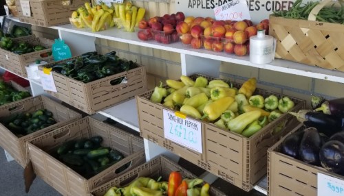 A display of fresh vegetables and fruits in wooden crates at a market, including peppers, cucumbers, and peaches.