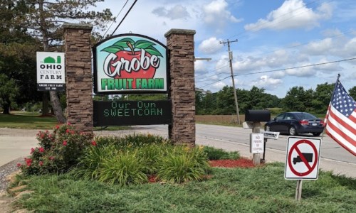 Sign for Grobe Fruit Farm featuring "Our Own Sweetcorn," with an American flag and nearby road signs.
