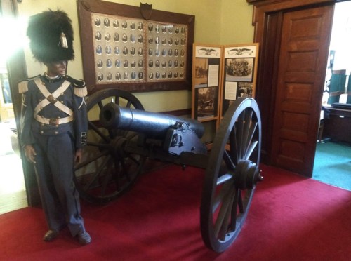 A historical display featuring a cannon and a soldier mannequin in a museum setting with framed photos on the wall.