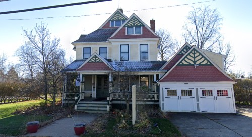A large, two-story Victorian-style house with a red roof, porch, and attached garage, surrounded by trees and grass.