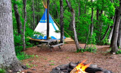 A campsite with a blue and white teepee, surrounded by trees, and a fire pit with a small fire in the foreground.