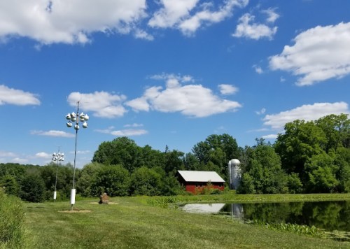 A serene landscape featuring a red barn, a silos, and a pond under a blue sky with fluffy clouds.