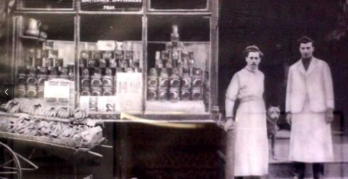 Two individuals stand outside a vintage shop displaying canned goods and other items in a historic setting.