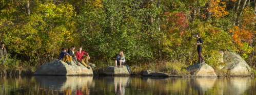 A group of people sitting on rocks by a calm lake, surrounded by vibrant autumn foliage.