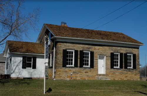 A stone house with a brown shingled roof, white door, and black shutters, set against a clear blue sky.