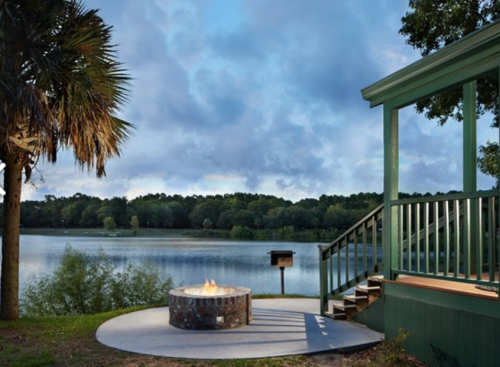 A serene lakeside scene featuring a fire pit, green deck, and lush trees under a cloudy sky.